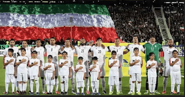 Members of Iran's national men's football team pose with chikdren as they sing their national anthem while waving their flag at the pitch in file photo. Iran World Cup ban in US imminent; FIFA sees Mexico as option